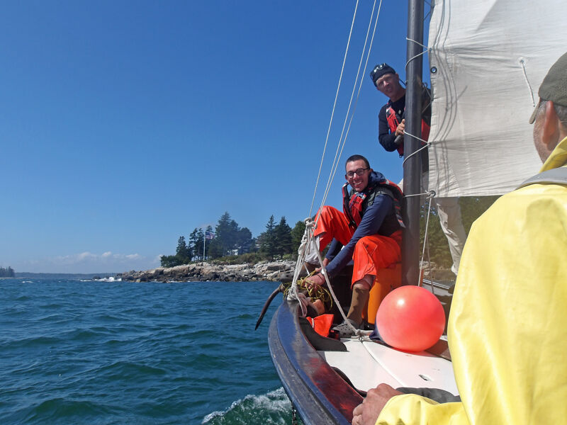 The image shows three people on a sailboat on a sunny day. The boat is sailing on a body of water, possibly the ocean, with land visible in the distance. One person is steering the boat, while the other two are sitting and standing on the deck. The boat has a white sail, and there is an orange buoy on the deck.